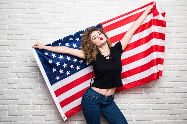 Beautiful young curly girl in casual clothes posing and smiling, standing covered with American flag against brick wall