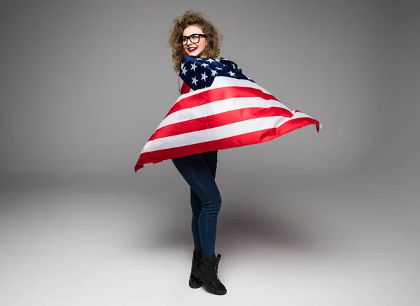 Веселая молодая женщина в повседневной одежде и очках покрыта американским флагом и улыбается на сером фоне — стоковое фото