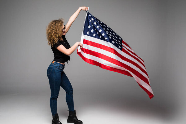 Cheerful young woman in casual clothes is wave the American flag and smiling on gray background