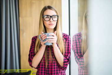 Genç bayan rahat kıyafetler içinde kahve içme ve bir pencereden bakarak serbest. Sabah.