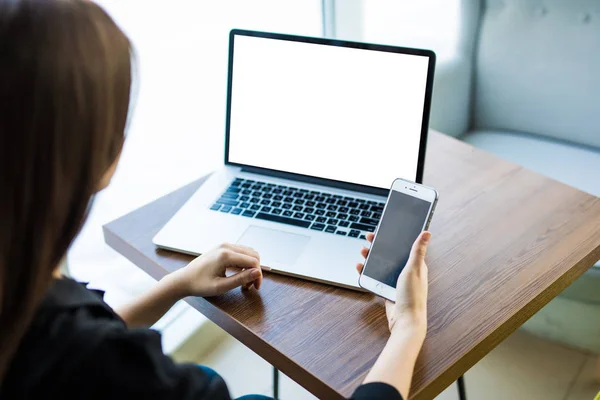 Vista superior, primer plano del teléfono inteligente con pantalla en blanco en manos de una joven sentada en la mesa redonda de madera con computadora portátil — Foto de Stock