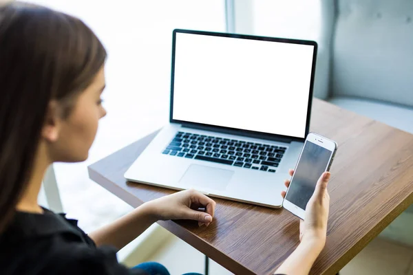 Vista superior, primer plano del teléfono inteligente con pantalla en blanco en manos de una joven sentada en la mesa redonda de madera con computadora portátil — Foto de Stock