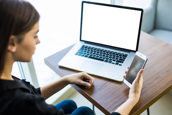 Vista superior, primer plano del teléfono inteligente con pantalla en blanco en manos de una joven sentada en la mesa redonda de madera con computadora portátil — Foto de Stock