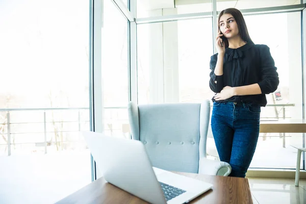 Mujer de negocios hablando por teléfono cerca de la mesa y mirando a la ventana — Foto de Stock