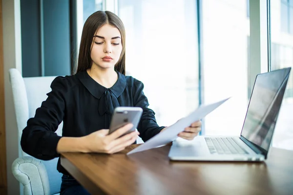 Joven mujer de negocios sentada en la mesa. Mujer firmar documentos, mantener el gráfico y el uso de teléfono inteligente. En la mesa está el ordenador portátil y la tableta. Estudiantes aprendiendo online . — Foto de Stock