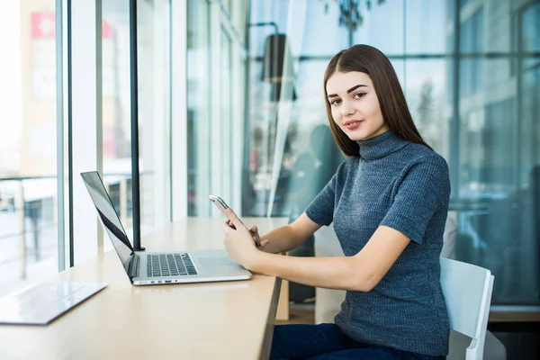 Vista lateral de la joven mujer de negocios sonriente sentado portátil en la mesa en la cafetería y utiliza el teléfono inteligente . — Foto de Stock