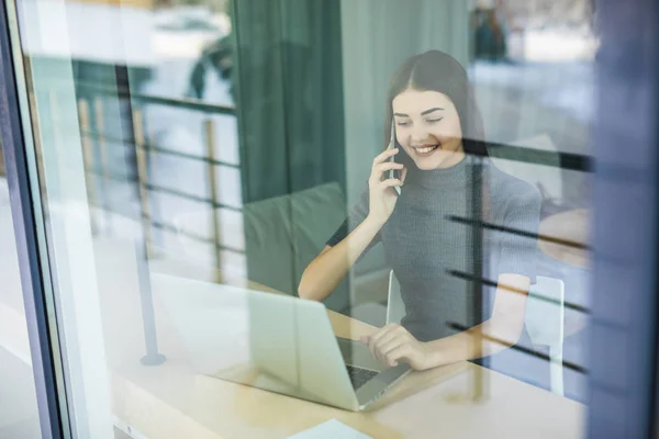 Mujer joven hablando por teléfono y utilizando la tableta a través de la ventana — Foto de Stock