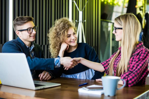 Los jóvenes estudiantes están usando aparatos, hablando y sonriendo mientras trabajan en la oficina moderna. Los hombres y la mujer están apretando las manos — Foto de Stock