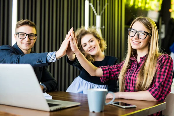 Teamwork en succes concept. Drie studenten juichen, geven hoge vijf aan elkaar, gelukkig na succesvolle presentatie van hun gemeenschappelijke project aan de Universiteit, zit op café met modern interieur — Stockfoto