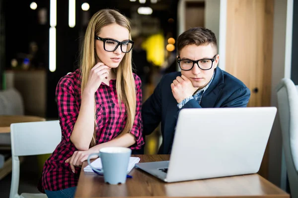 En ung, vakker kvinne som peker på en bærbar datamaskin med et smil og diskuterer noe med kollegaen sin mens hun står på kontoret – stockfoto