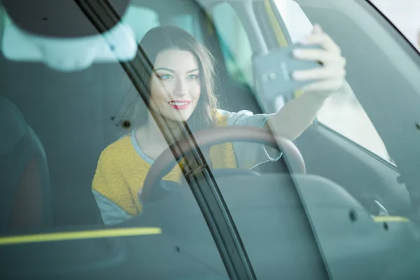 Улыбающаяся молодая женщина делает селфи с камерой смартфона в машине — стоковое фото