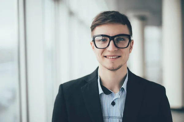 De cerca joven hombre de negocios sonriente con gafas, mirando a la cámara contra el pasillo de la oficina — Foto de Stock