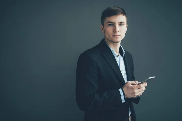 Hombre de negocios confiado apoyado contra una pared mientras usa el teléfono inteligente sobre fondo negro — Foto de Stock