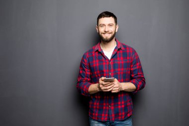 Kendine güvenen genç adam akıllı telefon tutarak ve gri arka plan karşı dururken kamera bakıyor