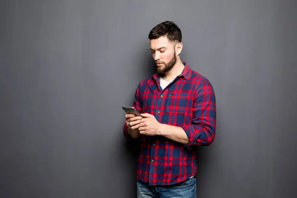 Kendine güvenen genç adam akıllı telefon tutarak ve gri arka plan karşı dururken bakmak — Stok fotoğraf