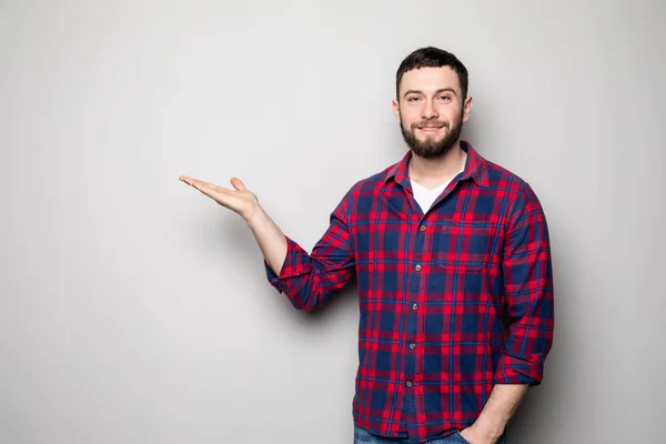 Attractive happy smiling casual young man in shirt holding copyspace on palm and showing okay gesture over gray background — Stock Photo, Image