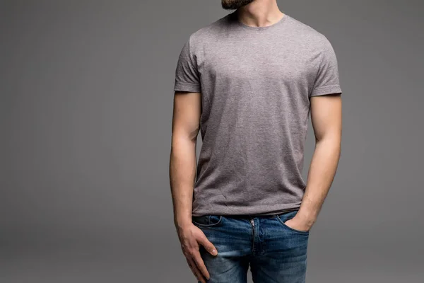 Un hombre con una camiseta gris y vaqueros sostiene sus manos en los bolsillos . —  Fotos de Stock