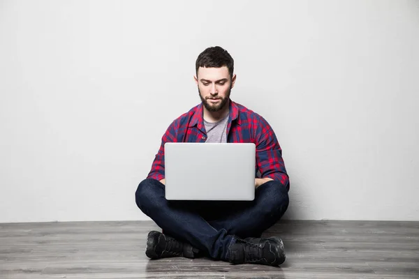 Jovem no interior com parede branca sentado no chão de madeira com laptop — Fotografia de Stock