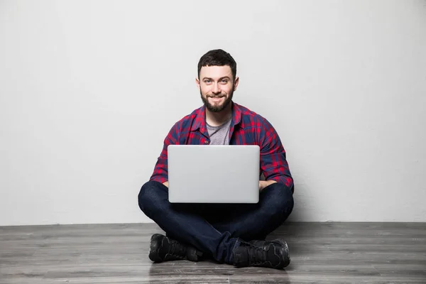 Молодой человек в интерьере с белой стеной сидит на деревянном полу с ноутбуком — стоковое фото
