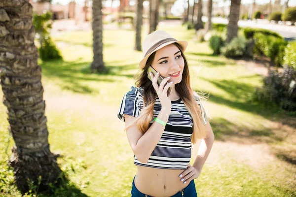 Smiling cute young woman in hat walking and talking on cell phone in summer resort. Summer time — Stock Photo, Image