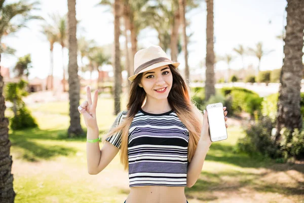 Femme avec signe de victoire tenir le téléphone avec écran blanc sur fond de paumes d'été — Photo