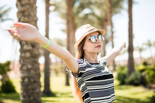 Souriant jeune femme détendue debout avec les mains levées dans le chapeau et les lunettes de soleil sur la station estivale — Photo