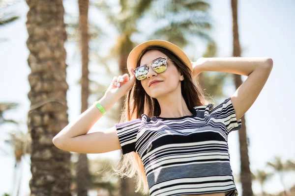 Mujer joven alegre en sombrero y gafas de sol manos sobre él en el fondo de las palmas de verano. Vocación de verano . —  Fotos de Stock