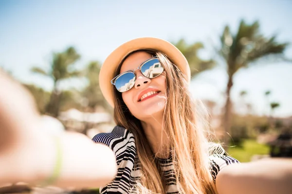 Sourire femme en chapeau et lunettes de soleil prenant selfie avec téléphone mobile des mains sur fond de palmiers station estivale. vocation estivale — Photo