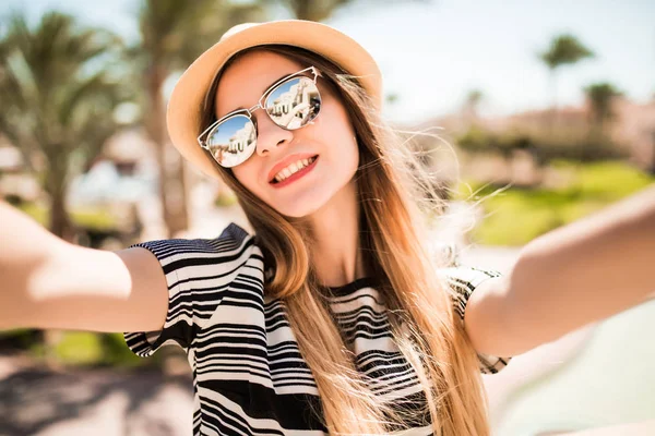 모자와 선글라스 여름 리조트 손바닥 배경에 손에서 휴대 전화와 함께 selfie를 복용에서 미소의 여자. 여름 직업 — 스톡 사진
