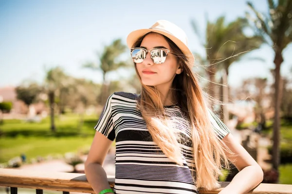 轻松的快乐微笑帽子和太阳镜的棕榈树背景上酒店的露台上的年轻女子的画像。暑假的时候 — 图库照片