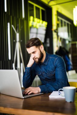 Dizüstü bilgisayarda çalışan ve kafede kahve tadını çıkarırken gülümseyen yakışıklı delikanlı
