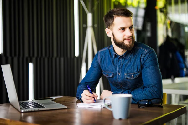 Joven guapo trabajando en el ordenador portátil y sonriendo mientras disfruta del café en la cafetería — Foto de Stock