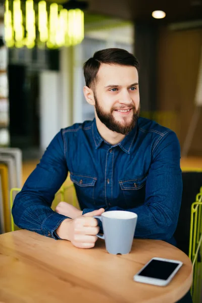 Guapo joven disfrutando de café en la cafetería mientras está sentado a la mesa con el teléfono tendido cerca de él — Foto de Stock