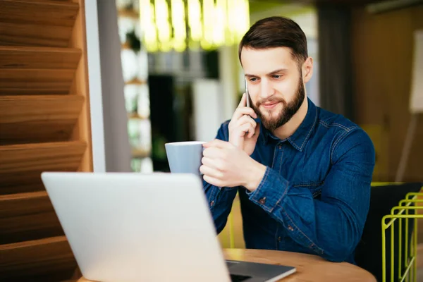 Homem bonito jovem com barba sentado no café falando telefone celular, segurando xícara de café e sorrindo. Laptop na mesa de madeira . — Fotografia de Stock