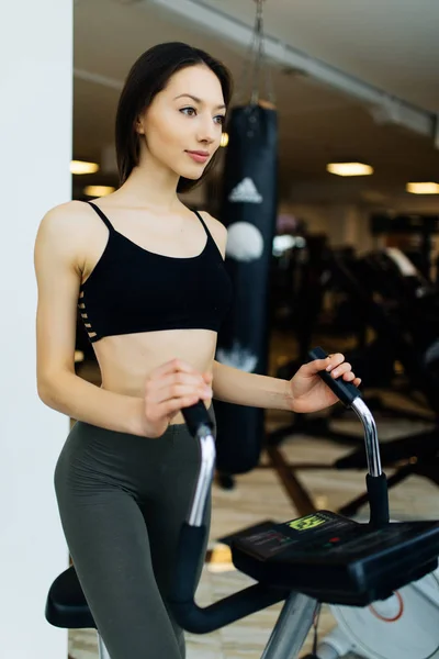 Mujer joven en forma usando un entrenador elíptico en un gimnasio y sonriendo. Retrato de chica fitness en el gimnasio cerca de una ventana, concepto de estilo de vida . — Foto de Stock