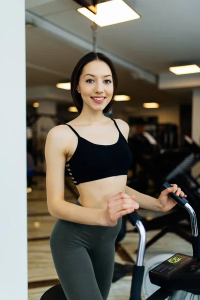 Mulher ajuste jovem usando um treinador elíptico em um centro de fitness e sorrindo. Retrato de menina fitness no ginásio perto de uma janela, conceito de estilo de vida . — Fotografia de Stock