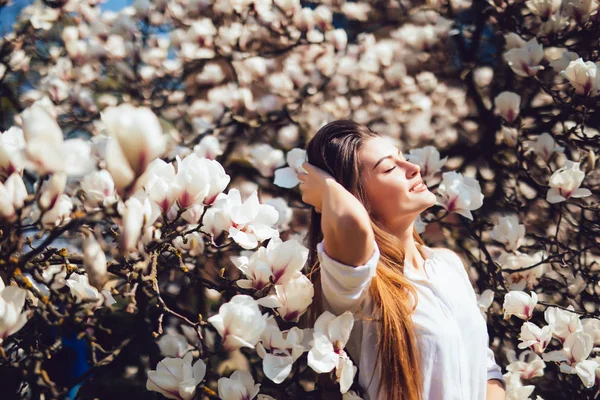 Ritratto all'aperto di una giovane bella donna vicino all'albero di magnolia con fiori ottenere bagno di sole. Ragazza che indossa vestiti eleganti. Umore primaverile femminile — Foto Stock