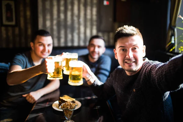 Três jovens em roupas casuais estão sorrindo, tomando selfie e bebendo cerveja enquanto estão sentados no pub — Fotografia de Stock