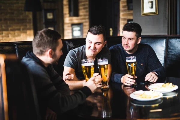 Tre unga män i casual kläder talar, äter chips och dricka öl när du sitter i pub och diskutera. — Stockfoto