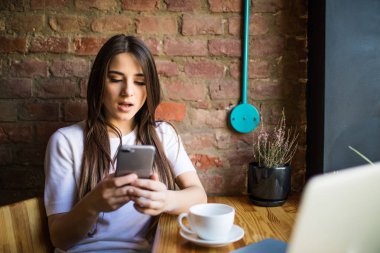 Kadın şok yazarak yazma iletide modern bir kafede akıllı telefon. Kahve ya da cep telefonuyla cappuccino bir masada oturan genç güzel kız Resim kırpılmış.