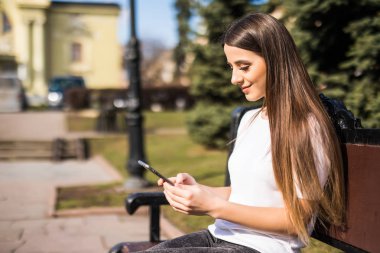 Bir bankta oturan bir şehirde bir akıllı telefon kullanan mutlu kız