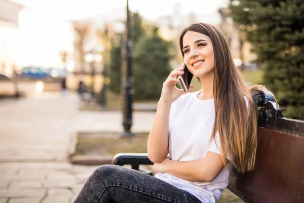 年轻漂亮的女人坐在长椅上在城市街道和谈电话和微笑 — 图库照片