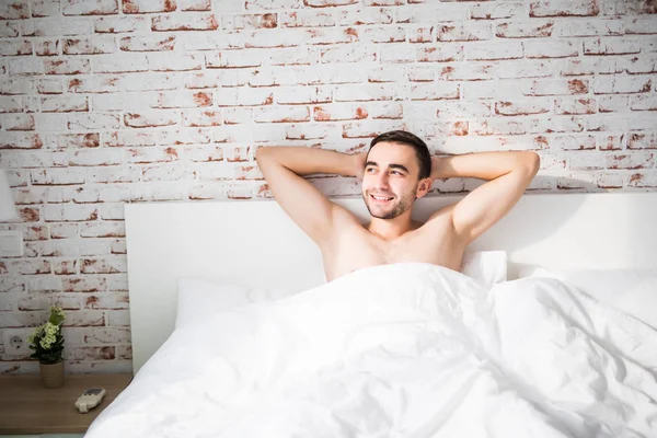Мечтательный мужчина расслабляется с руками над головой по утрам с улыбкой в спальне с кирпичной стеной — стоковое фото