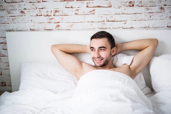 Красивый хорошо сложенный мужчина отдыхает в своей постели утром с улыбкой на кирпичной стене в комнате дома — стоковое фото