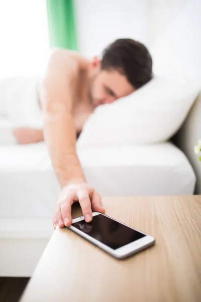 Красивый молодой человек просыпается с мобильным будильником утром в постели — стоковое фото