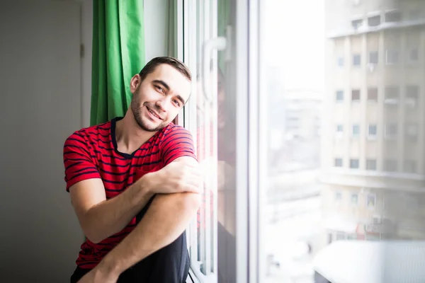 Genç adam onun apparment pencereye yakın oturan sabah Kameraya gülümse — Stok fotoğraf