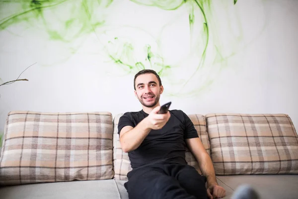 Bonito homem sorridente sentado em um sofá com um controle remoto em sua mão — Fotografia de Stock