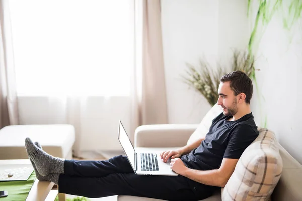 Hombre joven relajándose en el sofá con un ordenador portátil en casa — Foto de Stock