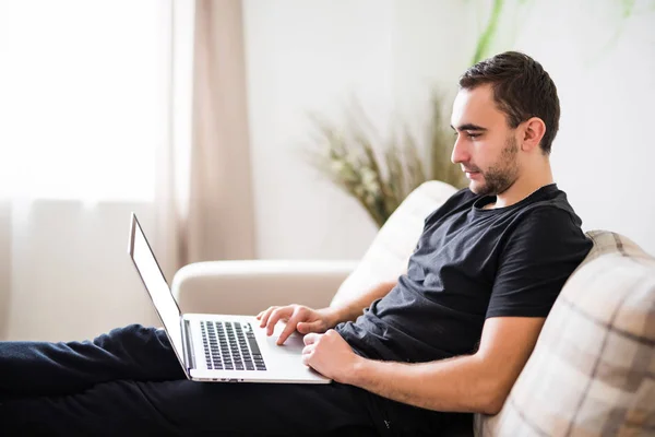 Vista lateral Hombre joven que se relaja en el sofá con un ordenador portátil en casa — Foto de Stock