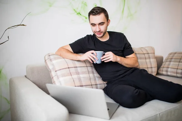 Guapo joven sonriente con taza de café usando el ordenador portátil en casa — Foto de Stock